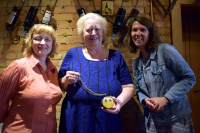 Three women standing in front of an interior brick wall. The woman in the center is holding a hockey puck decorated with gold glitter and googly eyes.